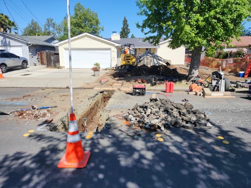Street excavation in front of a house with construction equipment, a trench, and orange traffic cones.