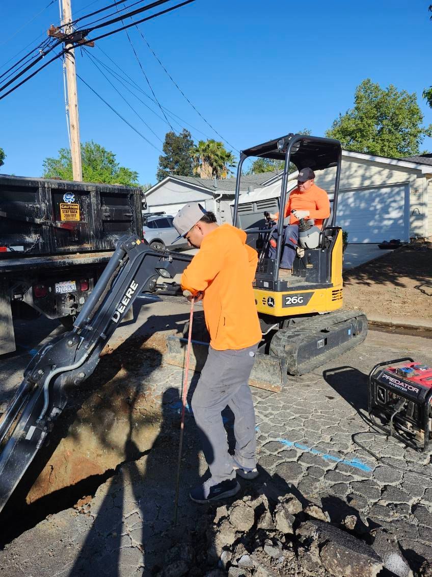 Construction workers operating an excavator next to a dump truck in a sunny, residential street. One worker is outside the machinery, inspecting something.