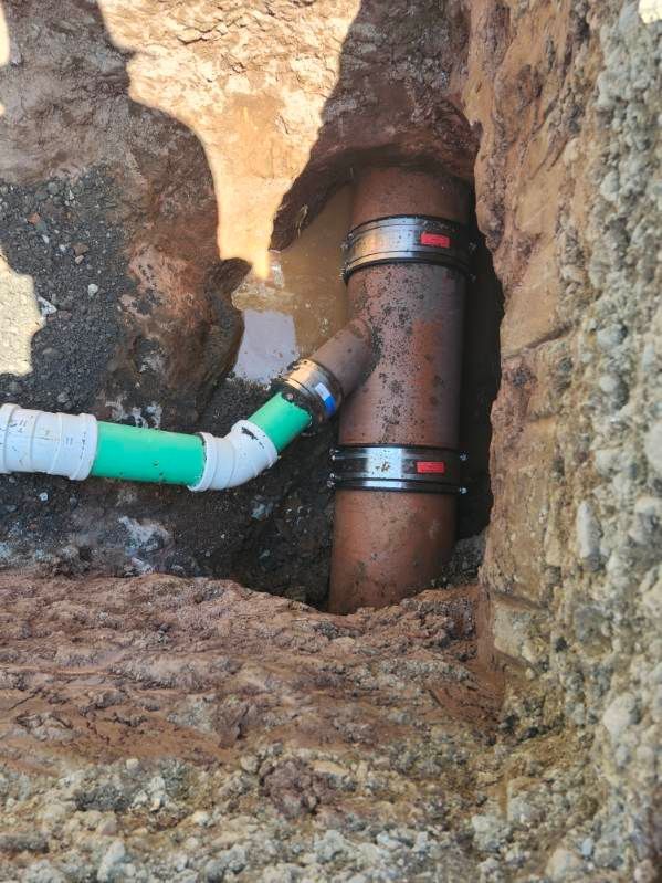 A plumbing repair in a dug trench. A brown pipe is joined by a smaller green and white pipe with large clamps securing the connections.