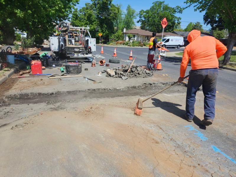 Workers in orange vests repair a street, raking asphalt and using equipment. White utility truck and cones set up for the work in a residential area.