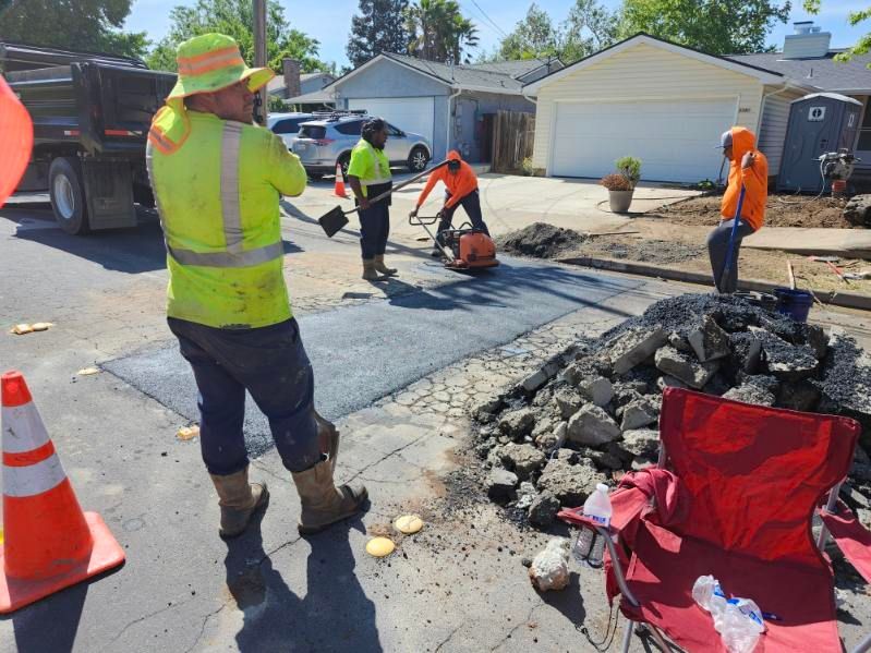 Road workers patching asphalt on a residential street. Men in safety vests and orange shirts operate machinery near a pile of debris.