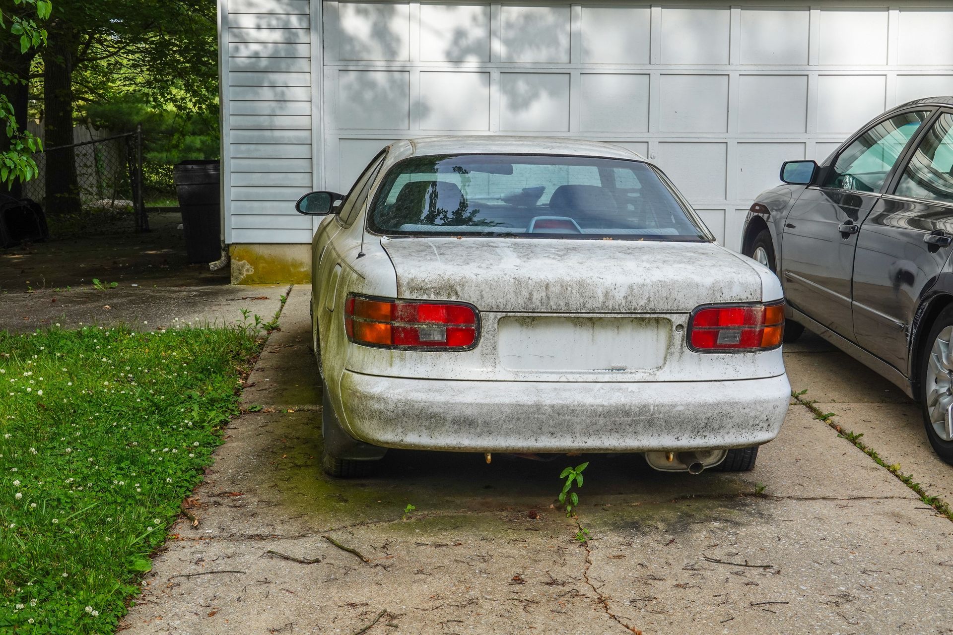an old, dirty white car parked on a driveway in front of a closed garage door.