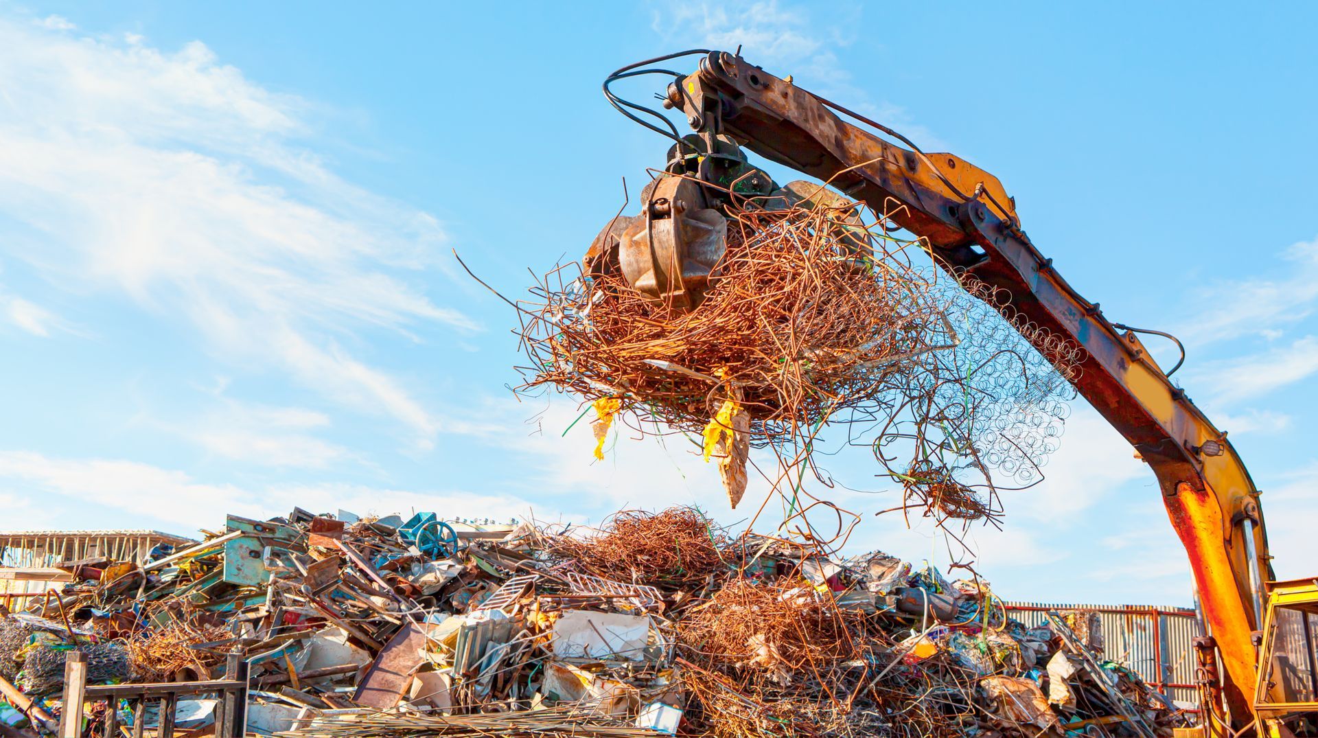 Excavator dumping crushed scrap metal at a recycling yard under a blue sky