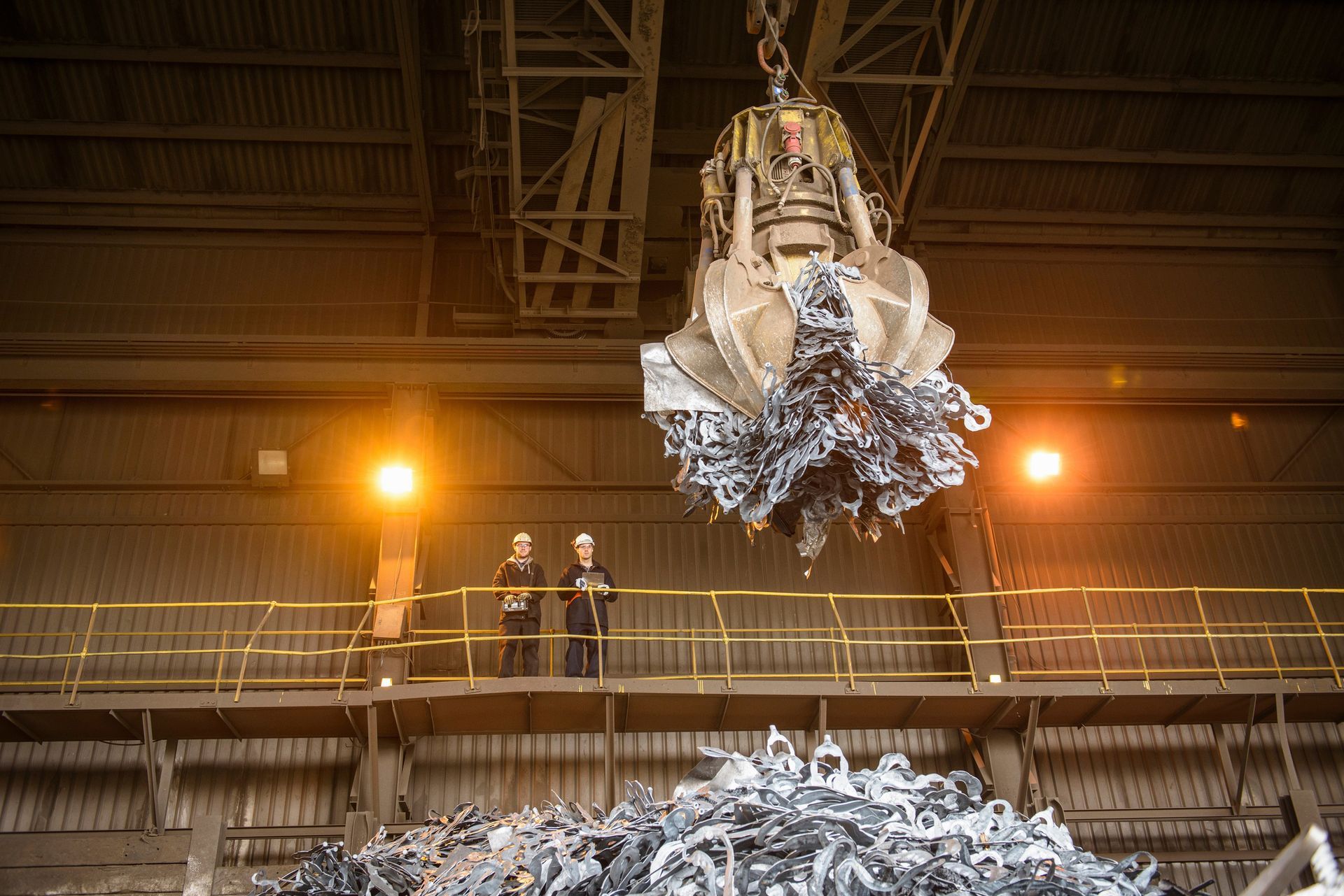 Two men wearing yellow protective helmets are watching a crane lift a pile of recyclable metal.