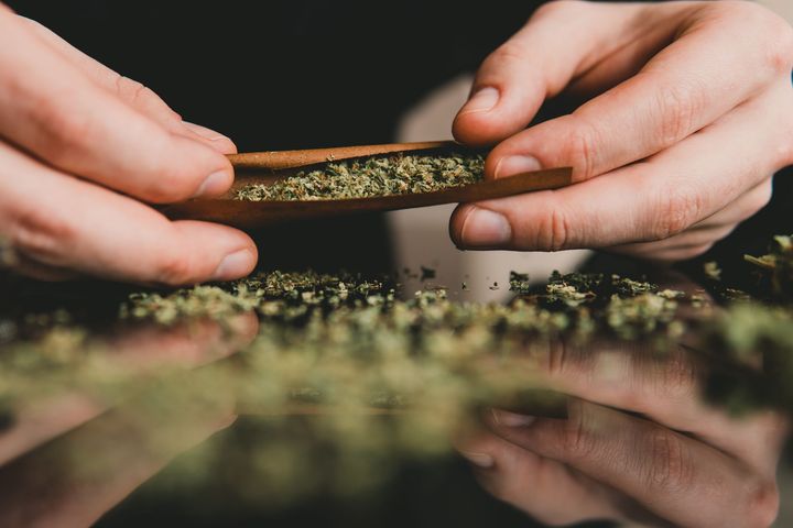 Hands rolling a cigar wrap filled with ground cannabis over a reflective surface.