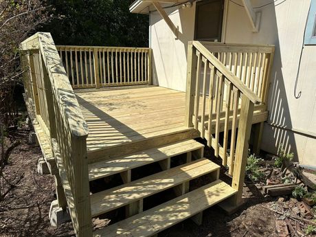 Wooden back deck with stairs and railing beside a house in a sunny yard.