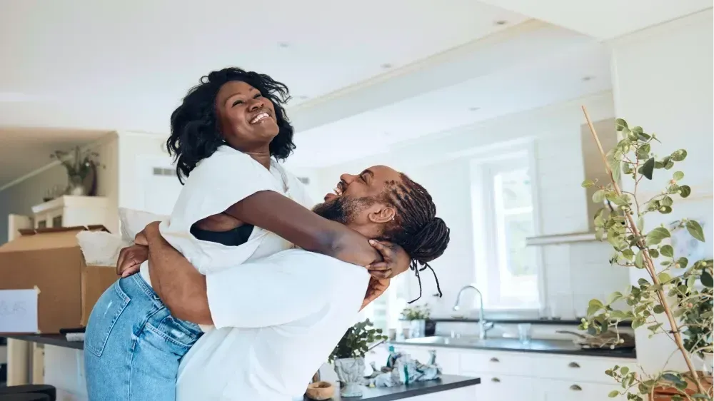 Couple embracing and laughing in a new home, boxes visible. White walls, kitchen counter, natural light.
