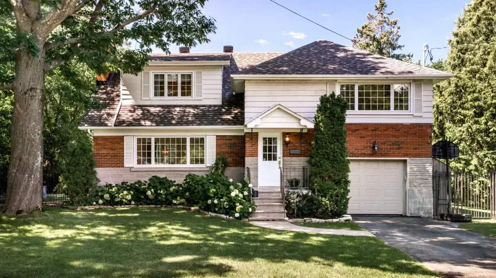 Two-story house with brick and white siding, front lawn, garage, and mature trees.