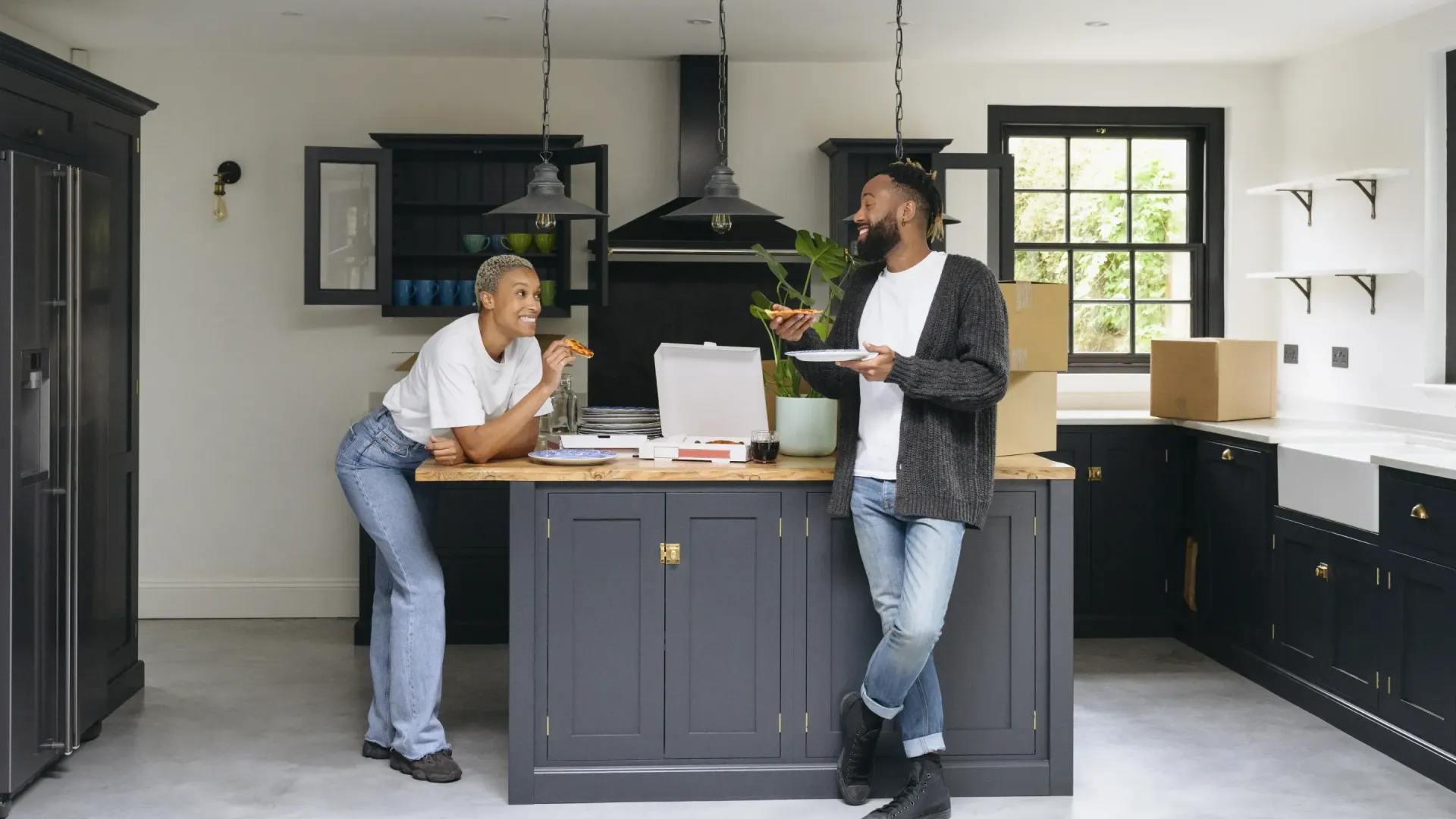 Couple in a new kitchen, smiling while eating pizza. Dark cabinetry, boxes, and white countertops are visible.