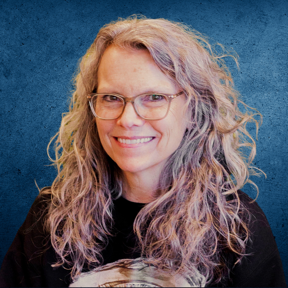 Woman with wavy, graying hair and glasses smiles against a blue background.