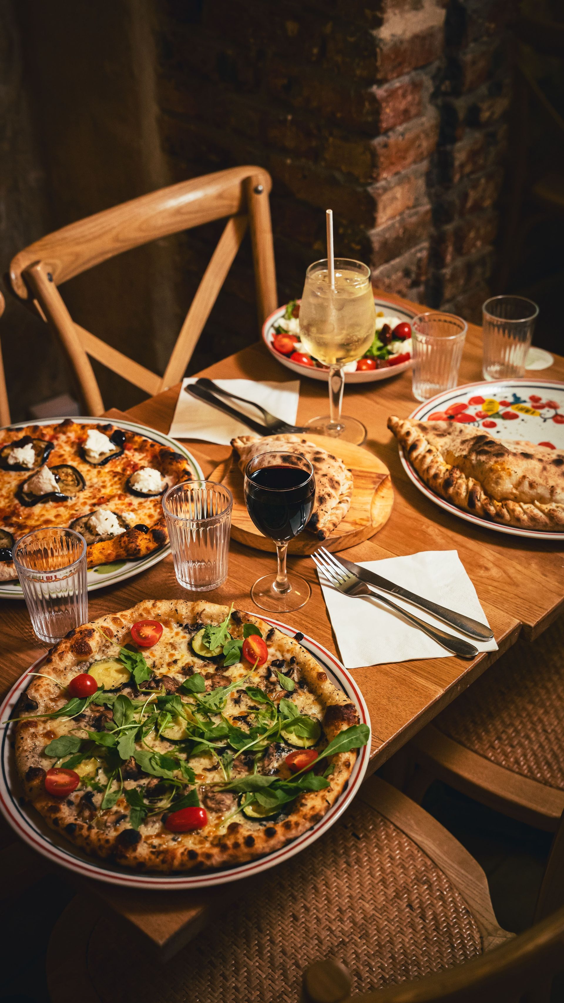 Pizza et vin sur une table en bois dans un restaurant. On aperçoit des assiettes, des verres et des chaises.