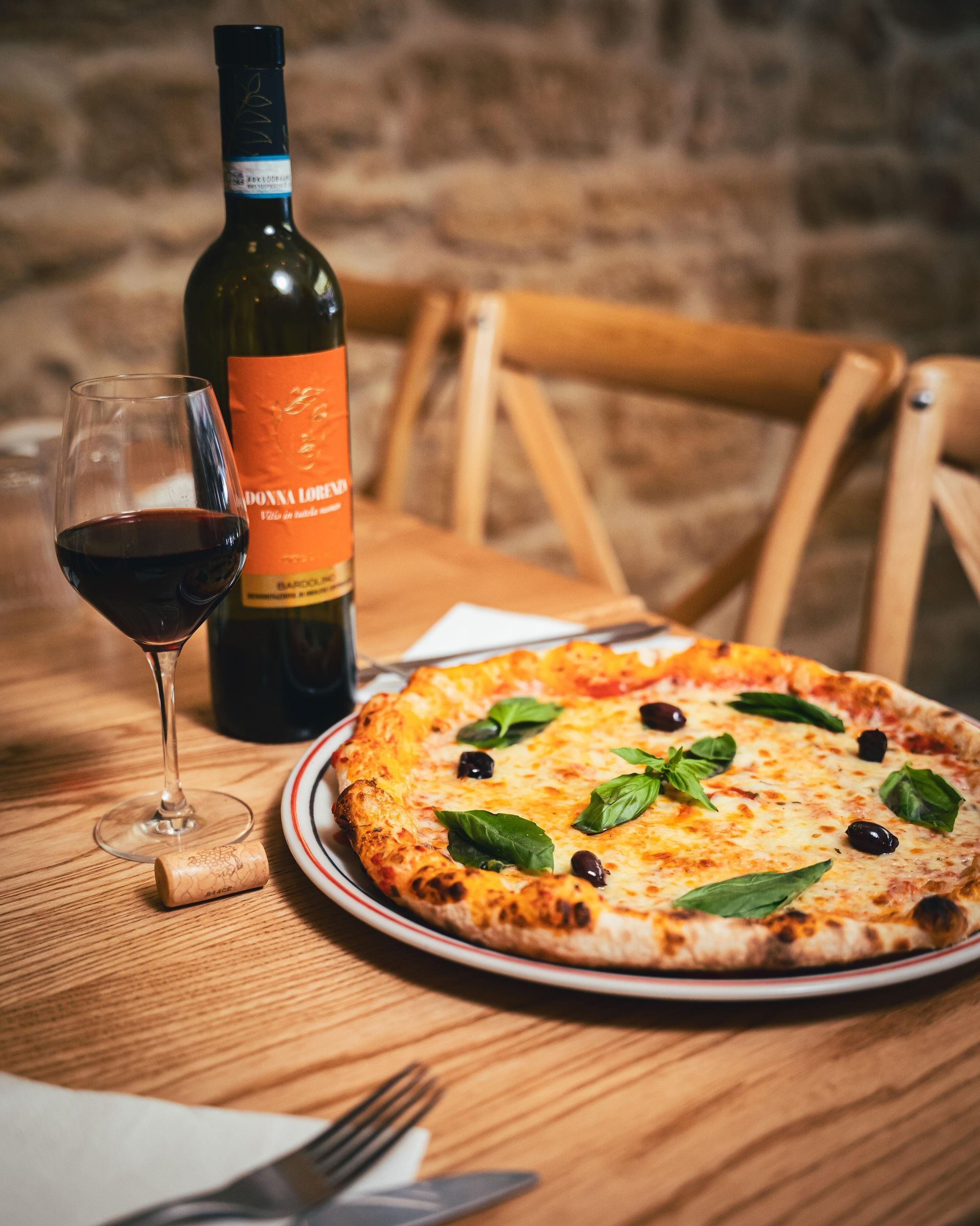 Pizza avec une bouteille de vin et un verre sur une table en bois. La pizza est garnie d'olives et de basilic.