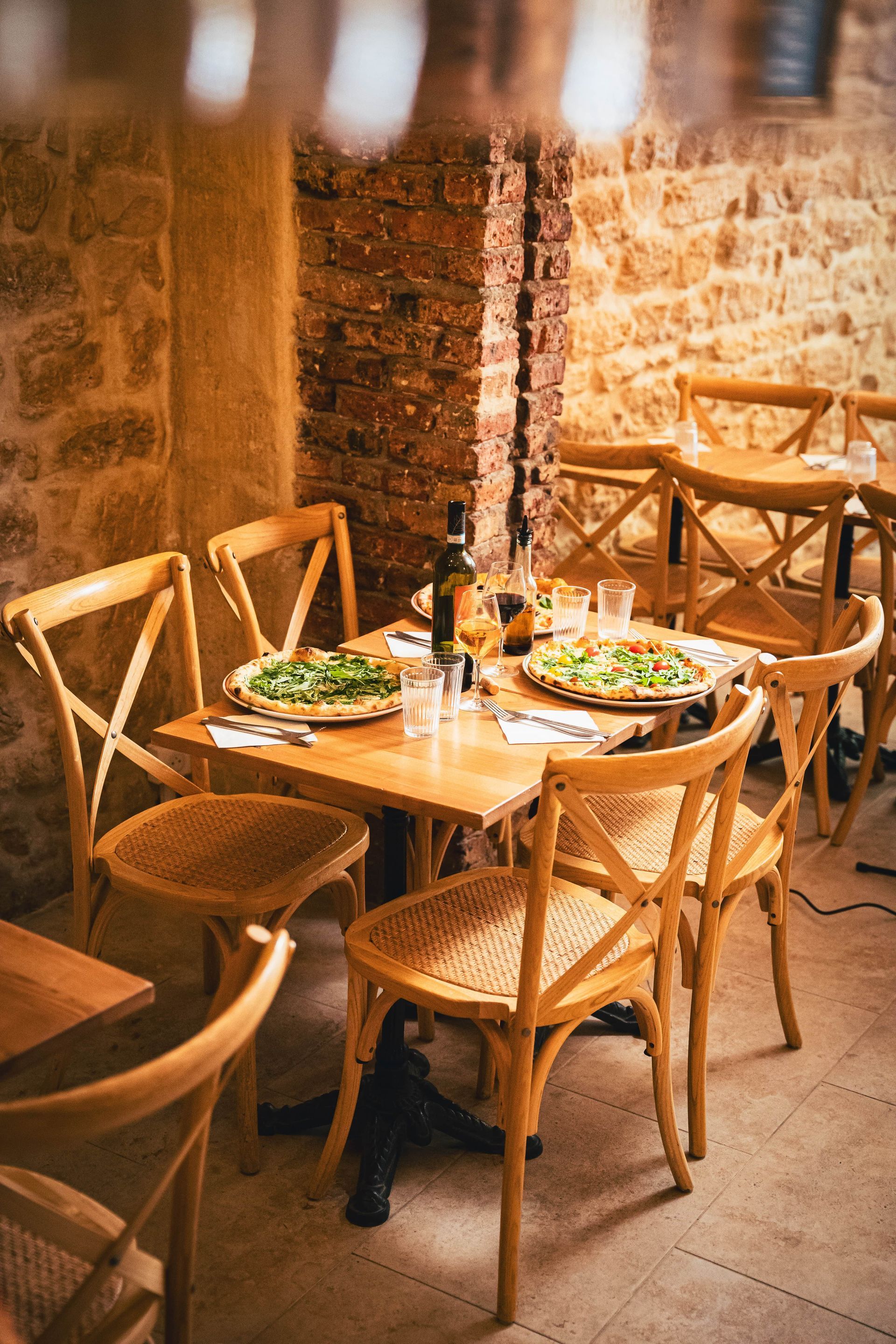Une table en bois est dressée pour un repas au restaurant. On aperçoit de la vaisselle, des verres et des chaises devant un mur en pierre.