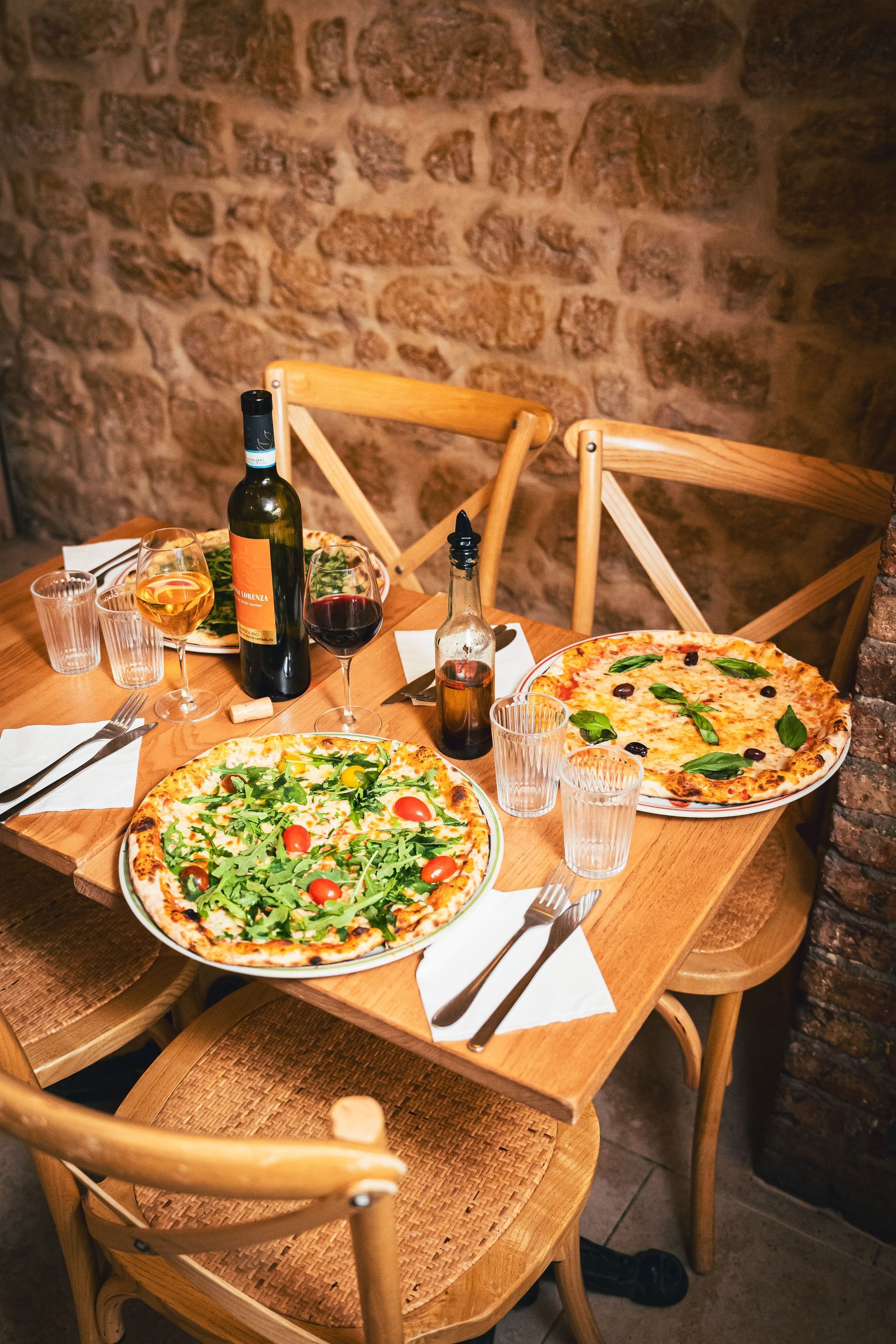 Une table en bois dressée avec des pizzas, des boissons et des couverts, entourée de chaises en bois, dans un cadre de restaurant rustique.