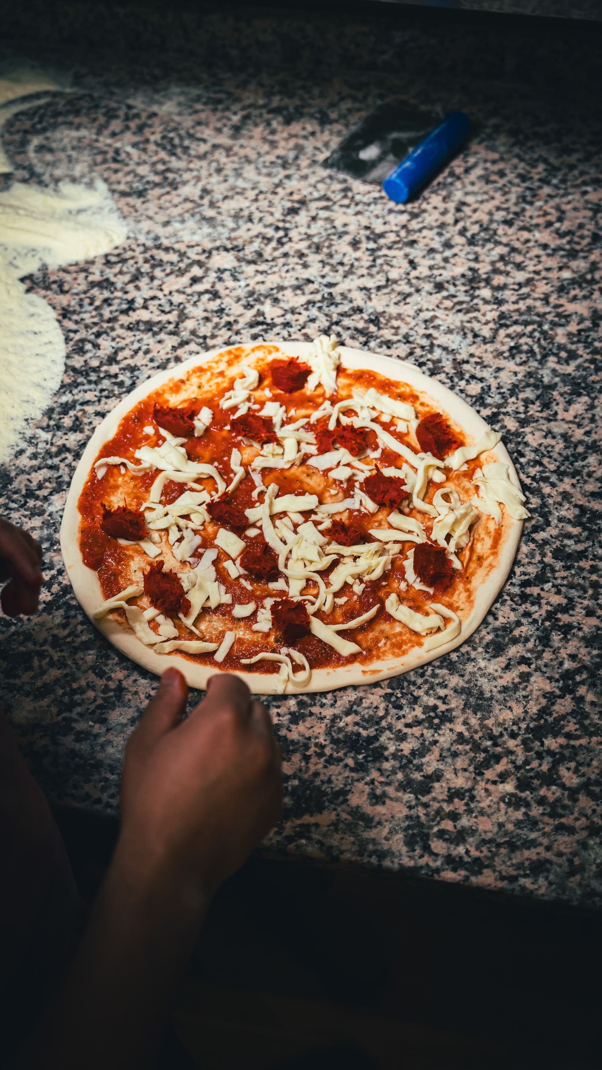 Préparation d'une pizza sur un plan de travail en granit : sauce, fromage et garnitures sont ajoutés.