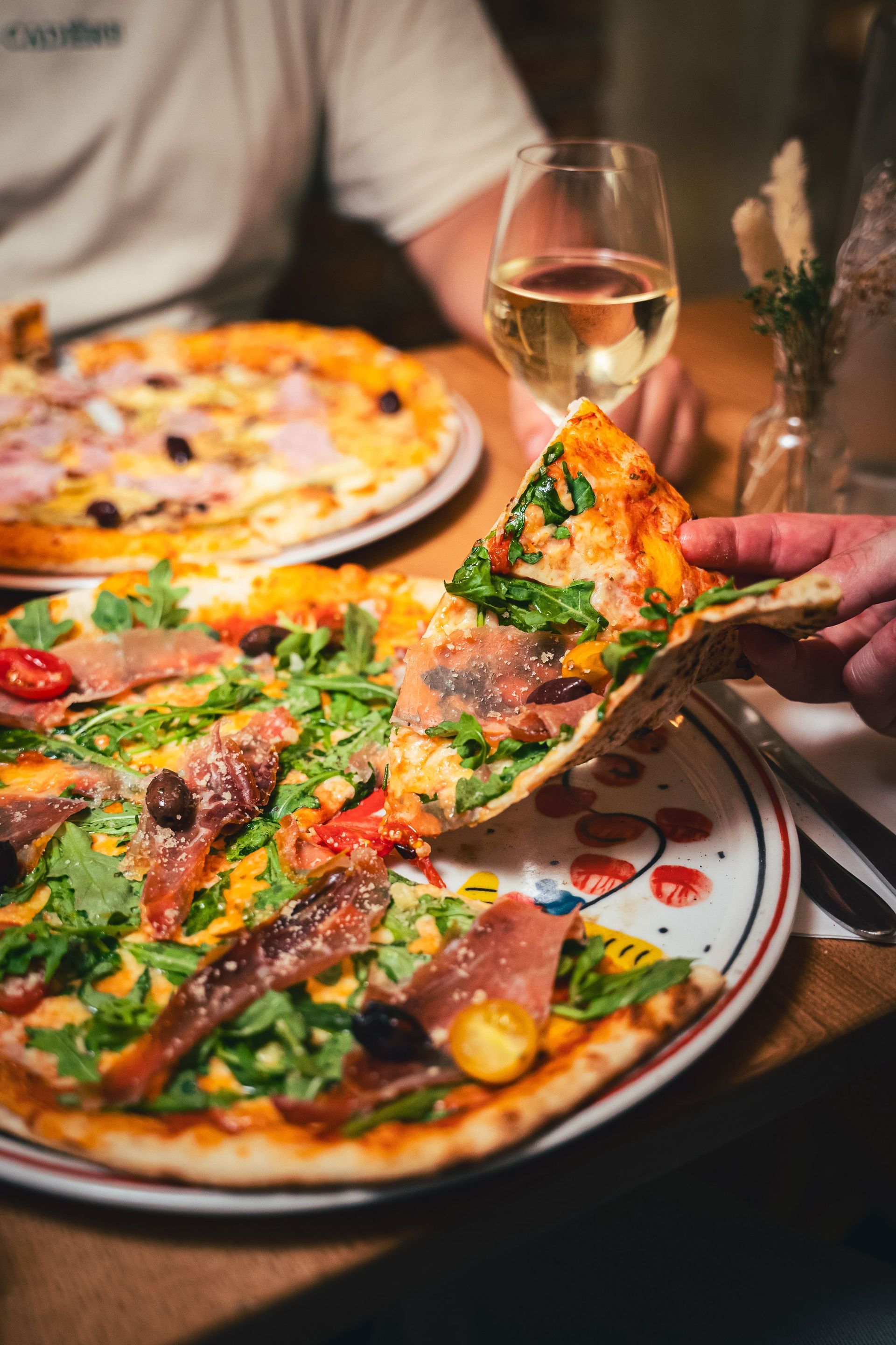 Une pizza garnie de prosciutto, de roquette et d'olives est retirée d'une assiette. Une personne tient un verre de vin blanc.