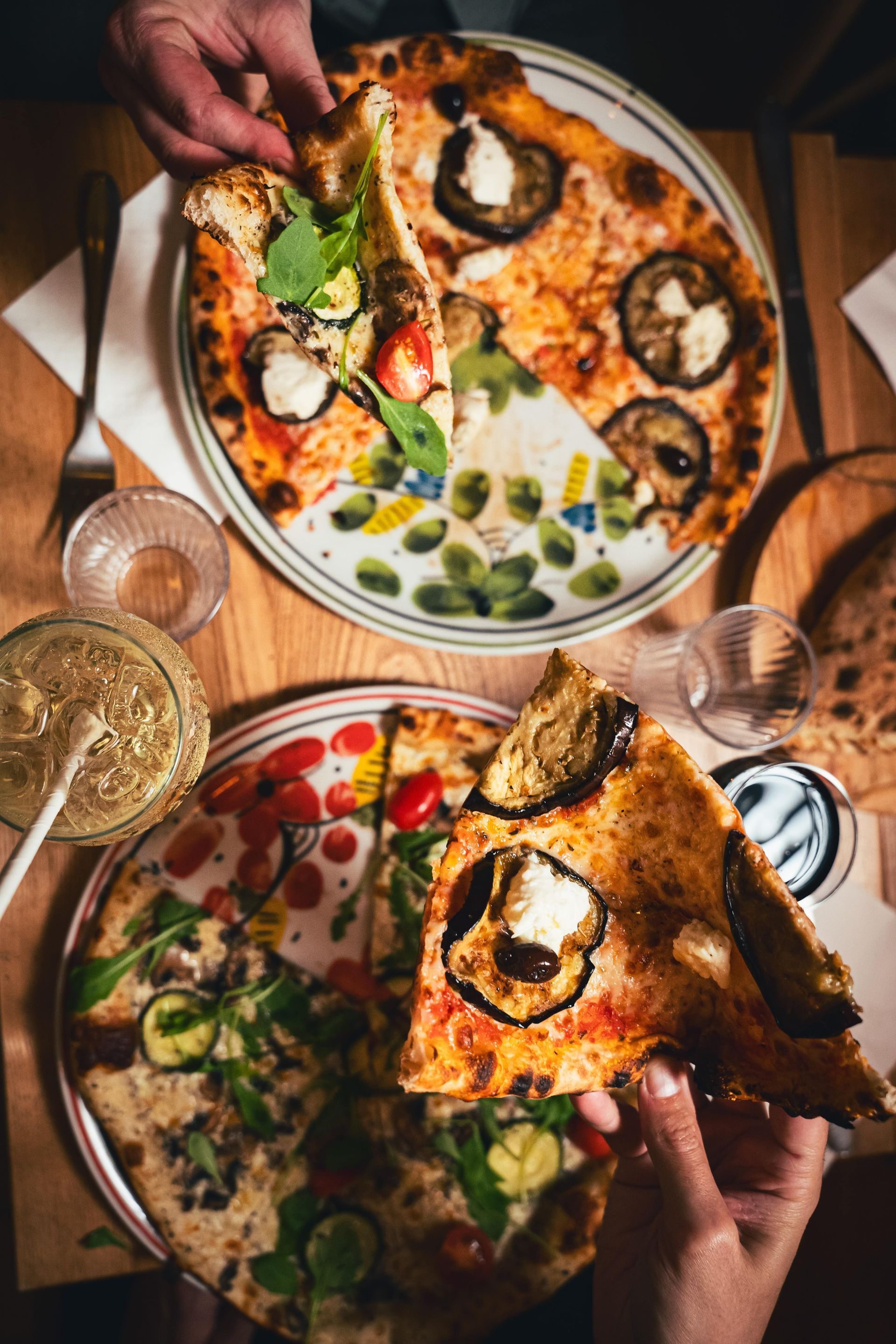 Deux pizzas sur des assiettes, en cours de service. Une part soulevée, table en bois, garnitures variées.