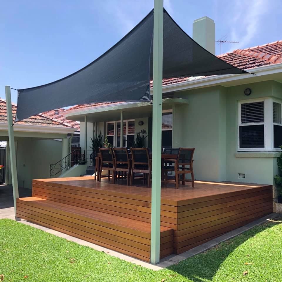 A wooden deck with a table and chairs under a shade sail.