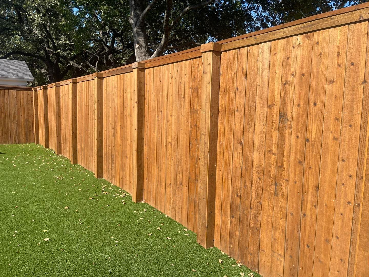 A row of vertical wooden fence panels with posts and a top trim, bordering a green lawn under a sunny sky.