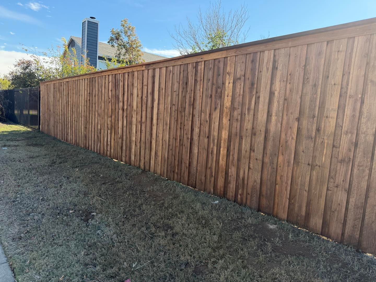 A long, stained wooden privacy fence stretches across a sunny residential backyard with sparse grass and a house in back.