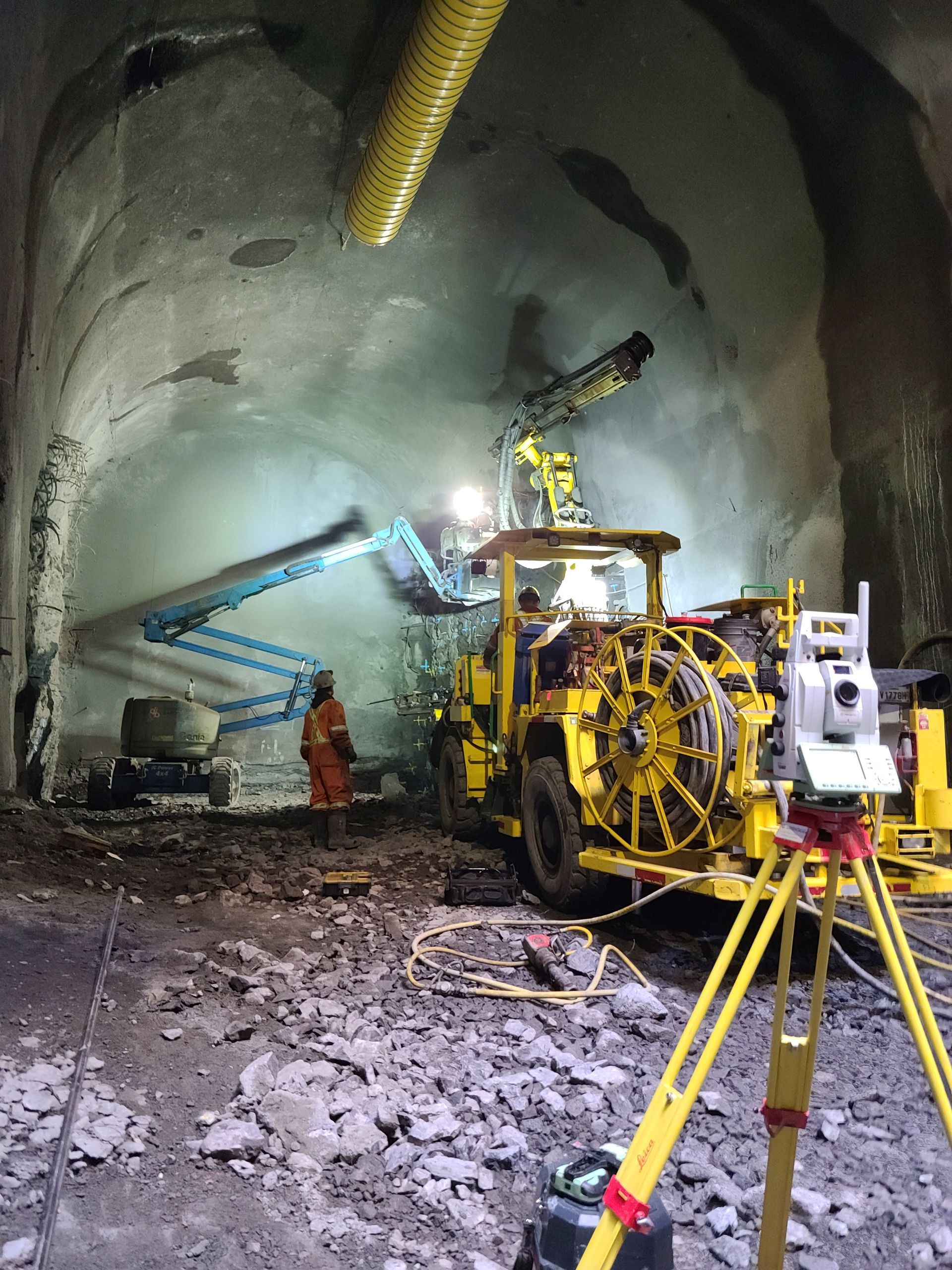 Un ouvrier et des engins lourds, dont une foreuse jaune et un élévateur bleu, sont à l'œuvre à l'intérieur d'un tunnel en construction faiblement éclairé.