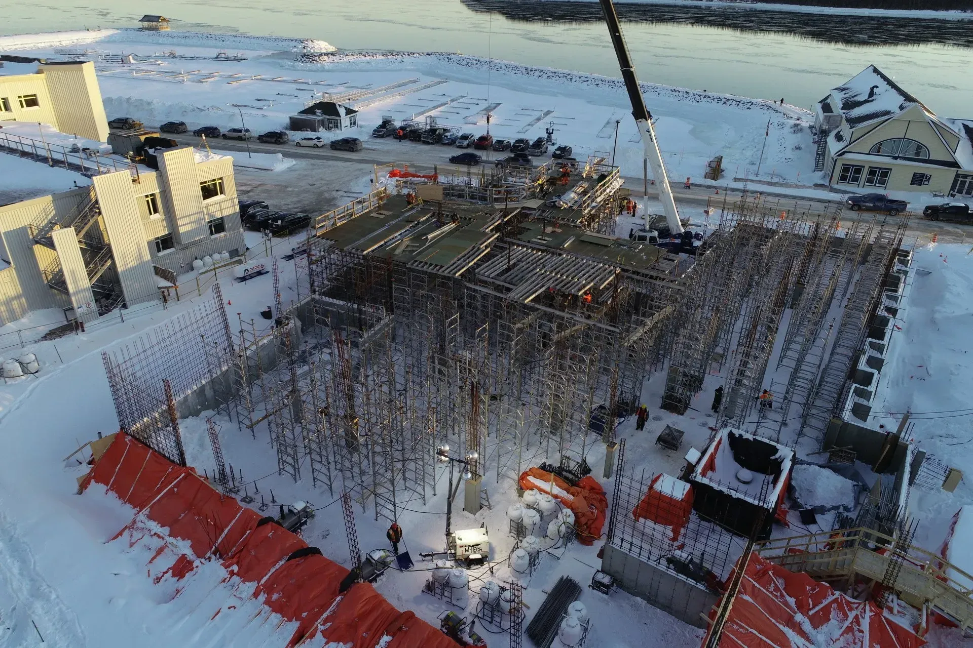 Une vue aérienne montre un chantier enneigé avec une structure métallique, une grue et des bâches de protection orange.