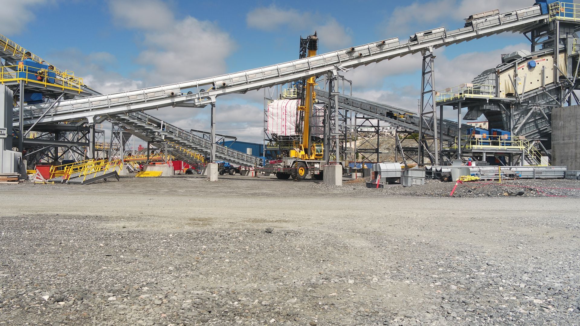 Site minier ou de construction industriel avec de hauts convoyeurs, des structures métalliques et une grue sous un ciel nuageux.