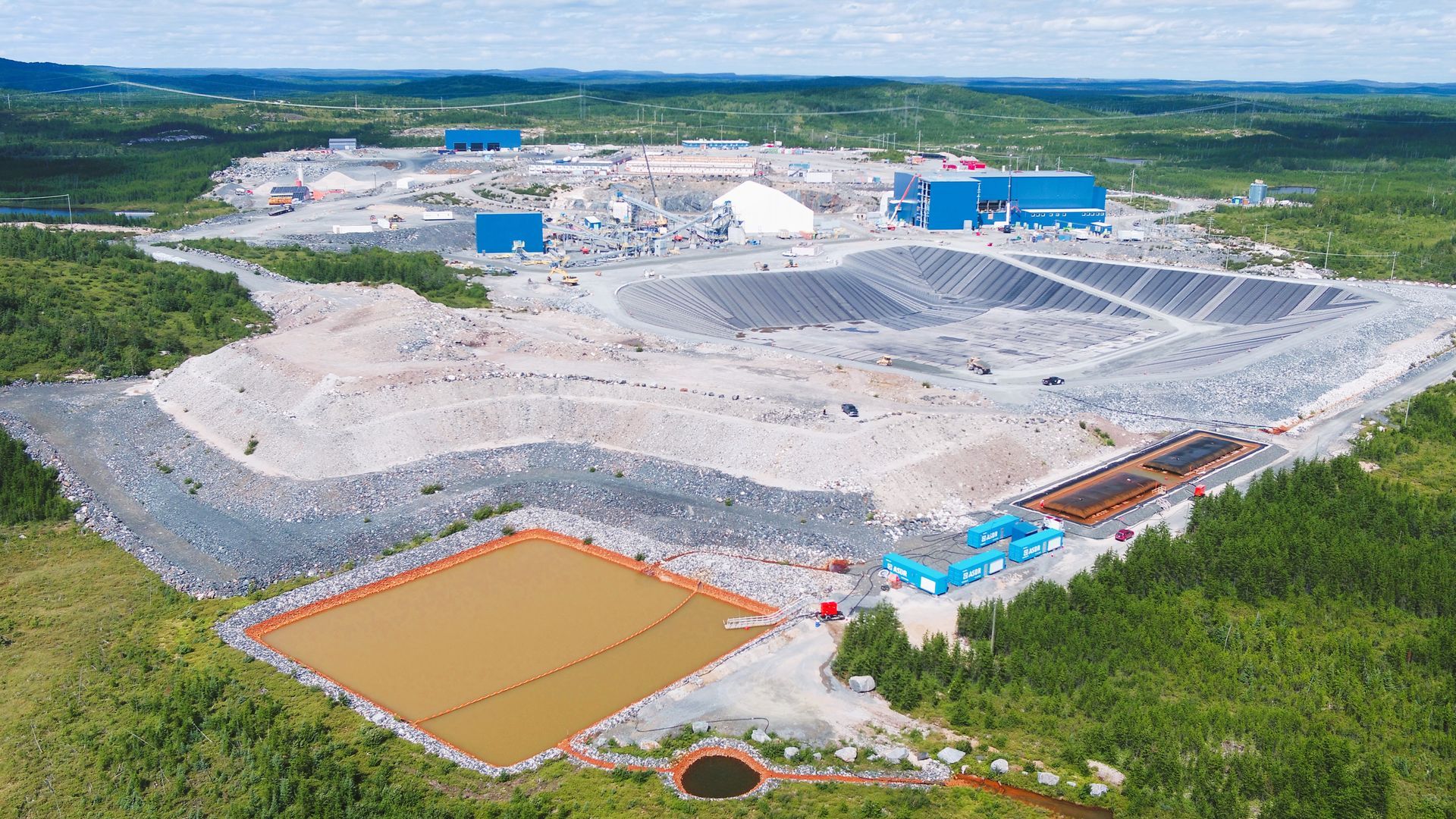 Vue aérienne d'un site minier industriel avec des bâtiments bleus, de grands tas de roches et un bassin de décantation carré de couleur beige.