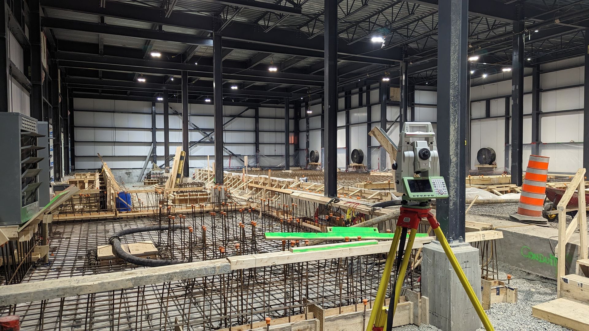 Une station totale de géomètre, montée sur un trépied, se trouve à l'intérieur d'un grand entrepôt en construction, renforcé par une grille métallique.