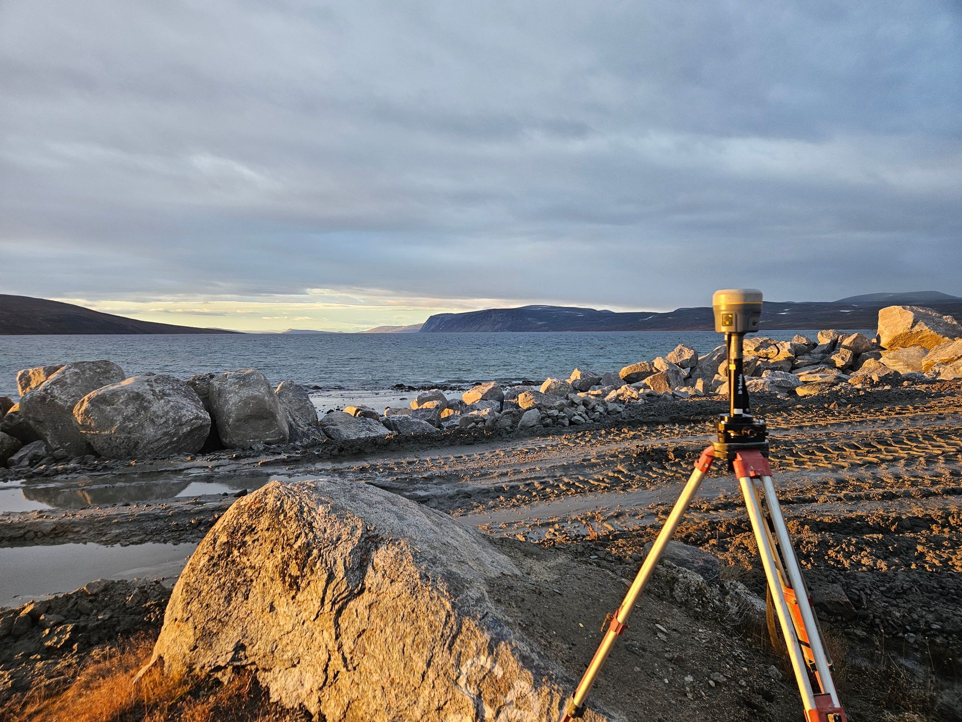 Un récepteur GNSS de topographie, monté sur un trépied, se dresse sur un rivage rocheux au bord d'un grand lac, sous un ciel nuageux de fin d'après-midi.