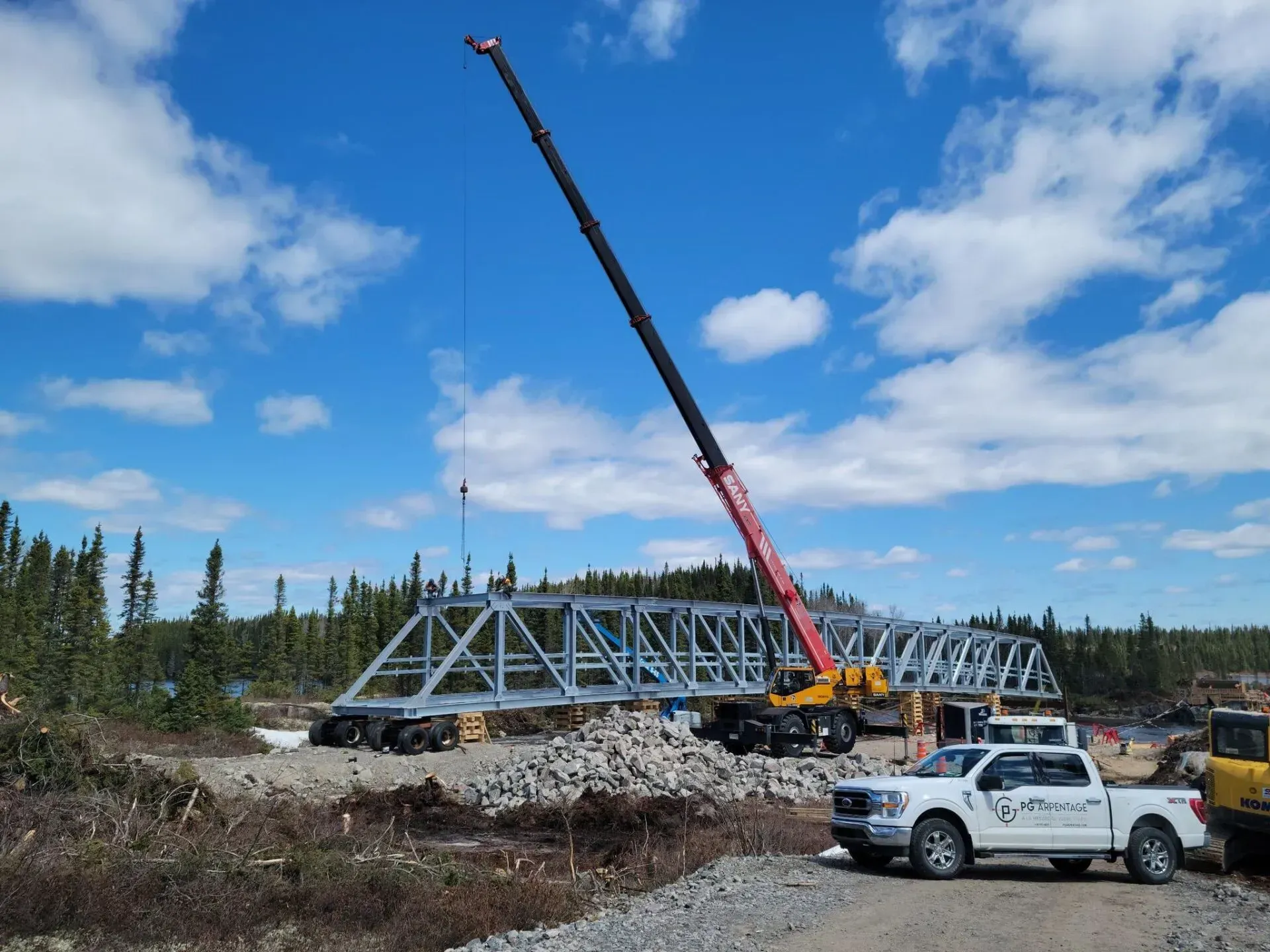 Une grue soulève un grand tronçon de pont métallique sur un chantier de construction entouré d'arbres, sous un ciel d'un bleu éclatant.
