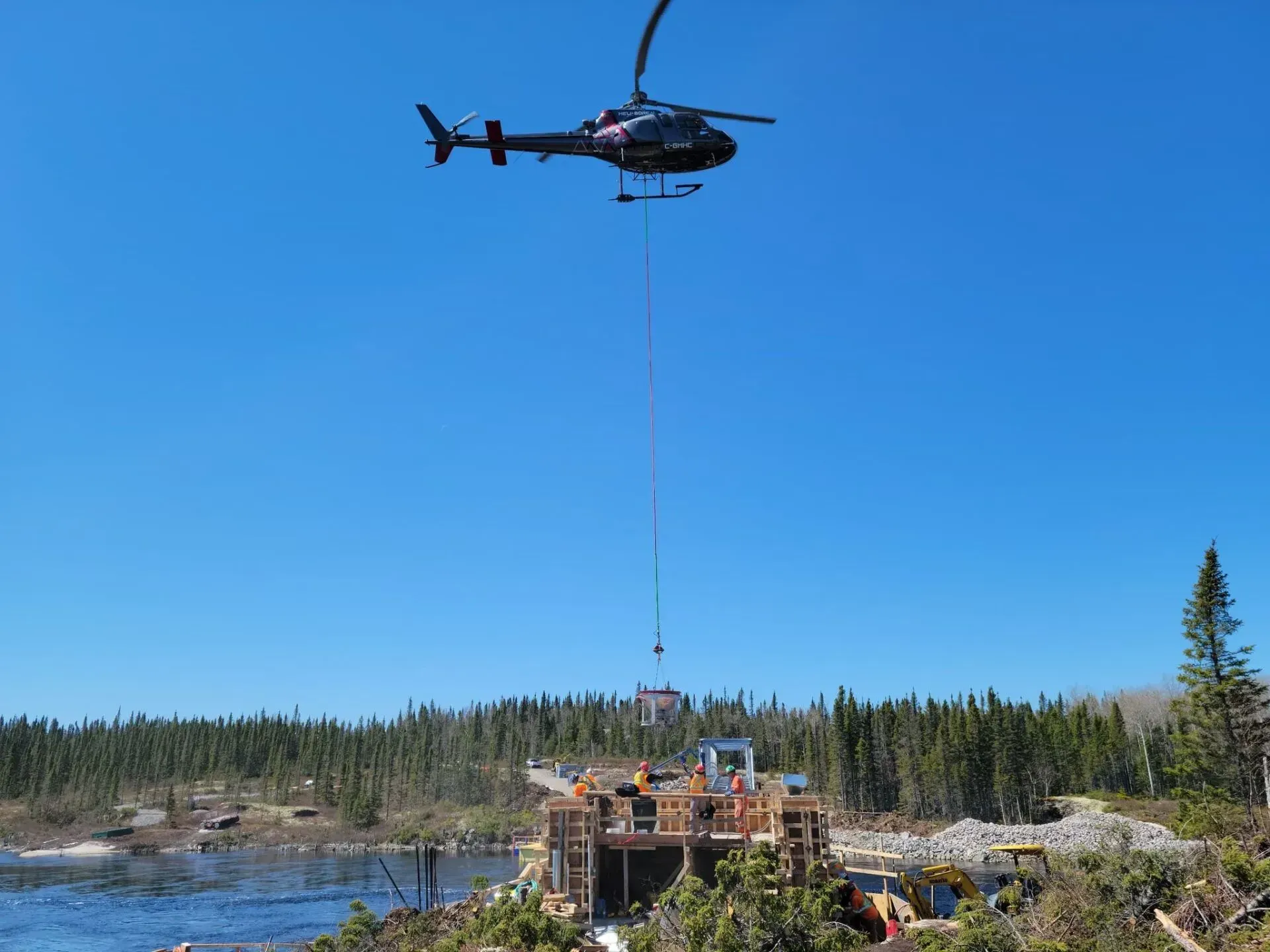 Un hélicoptère survole un chantier de construction près d'un plan d'eau, descendant des matériaux de construction à l'aide d'un long câble.