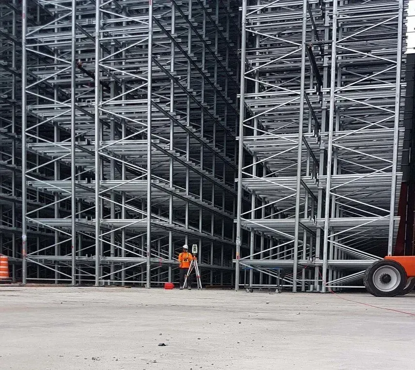 Un ouvrier portant un gilet orange haute visibilité utilise un trépied d'arpenteur devant de grands rayonnages métalliques industriels à plusieurs niveaux.