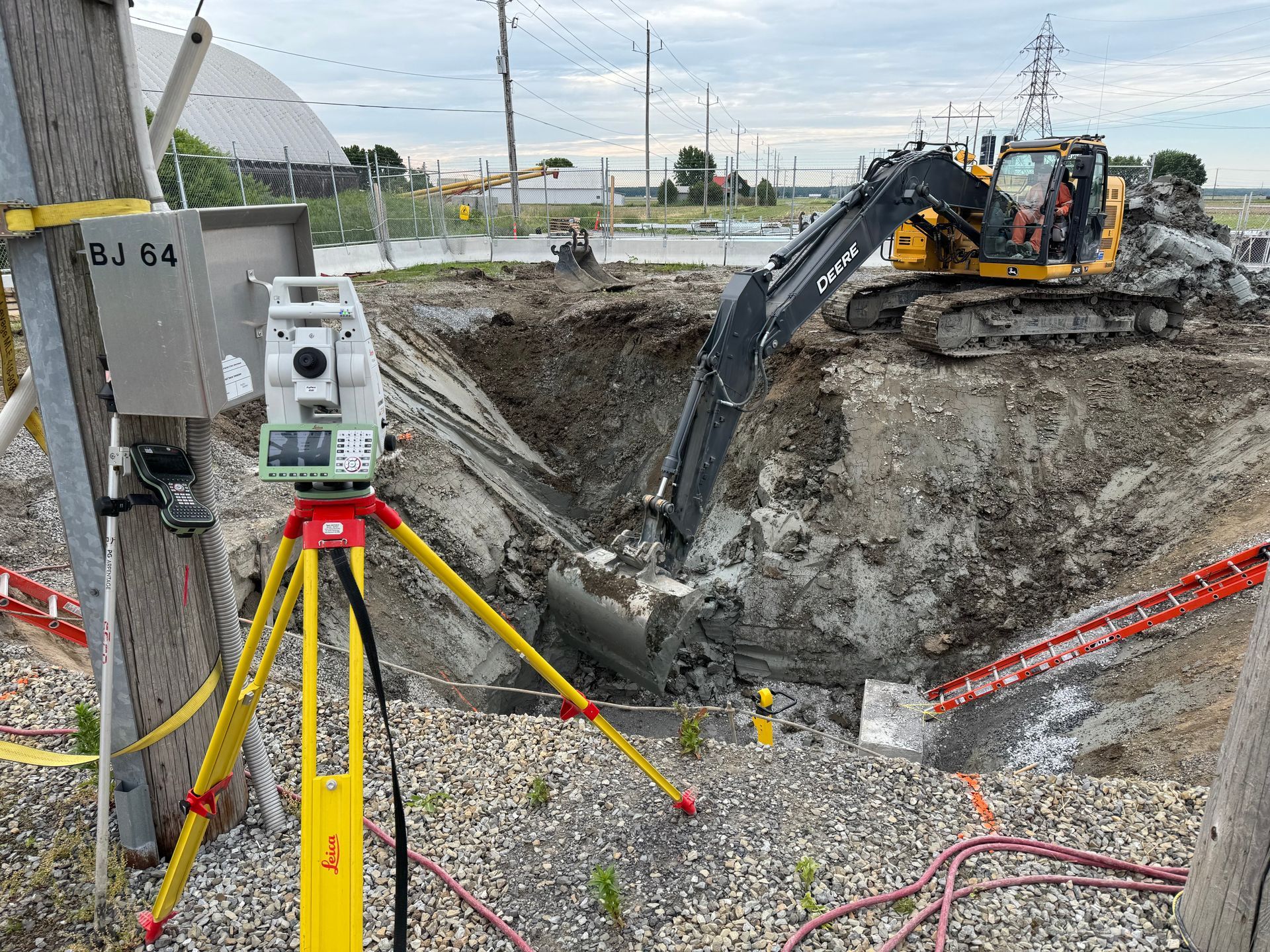 Une pelleteuse jaune creuse un chantier de construction en terre, vue à travers un instrument de topographie sur trépied.