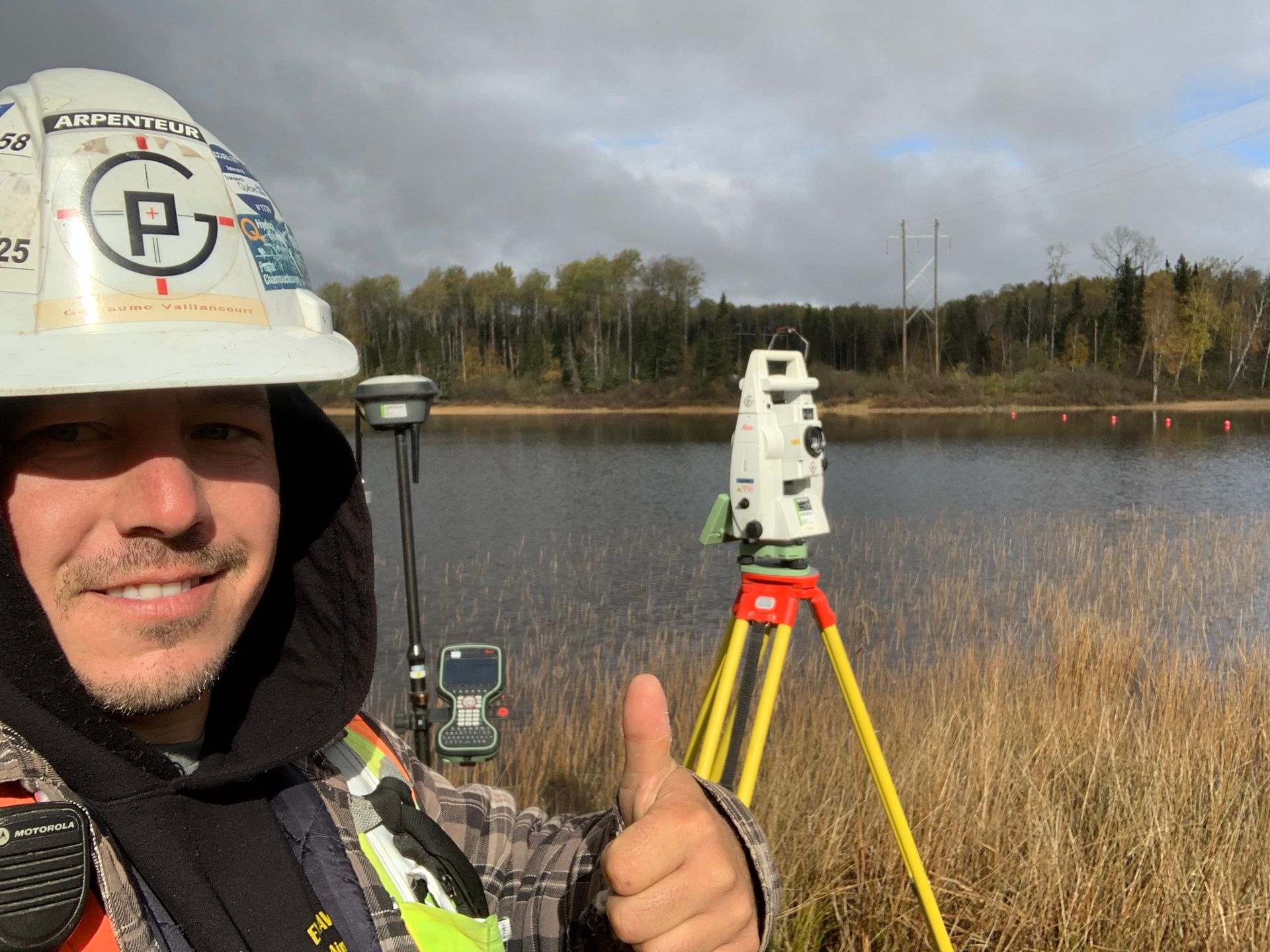 Un géomètre portant un casque de chantier lève le pouce à côté de son matériel de topographie au bord d'un lac.