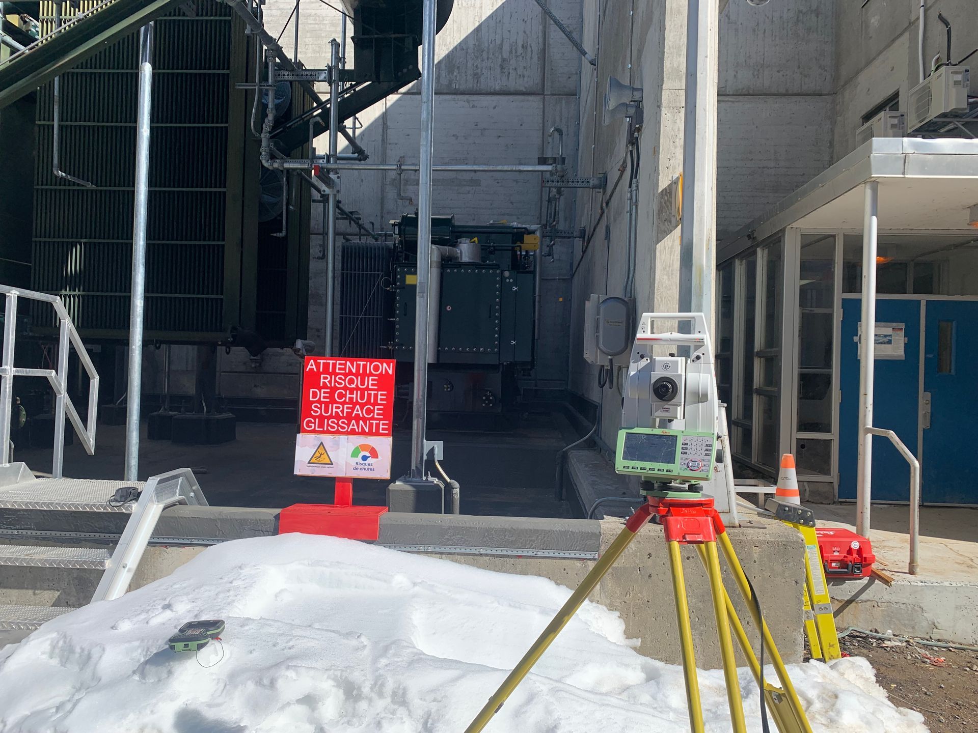 Un instrument d'arpentage sur trépied se dresse près d'un sol enneigé, un panneau d'avertissement étant apposé sur une petite barrière rouge.
