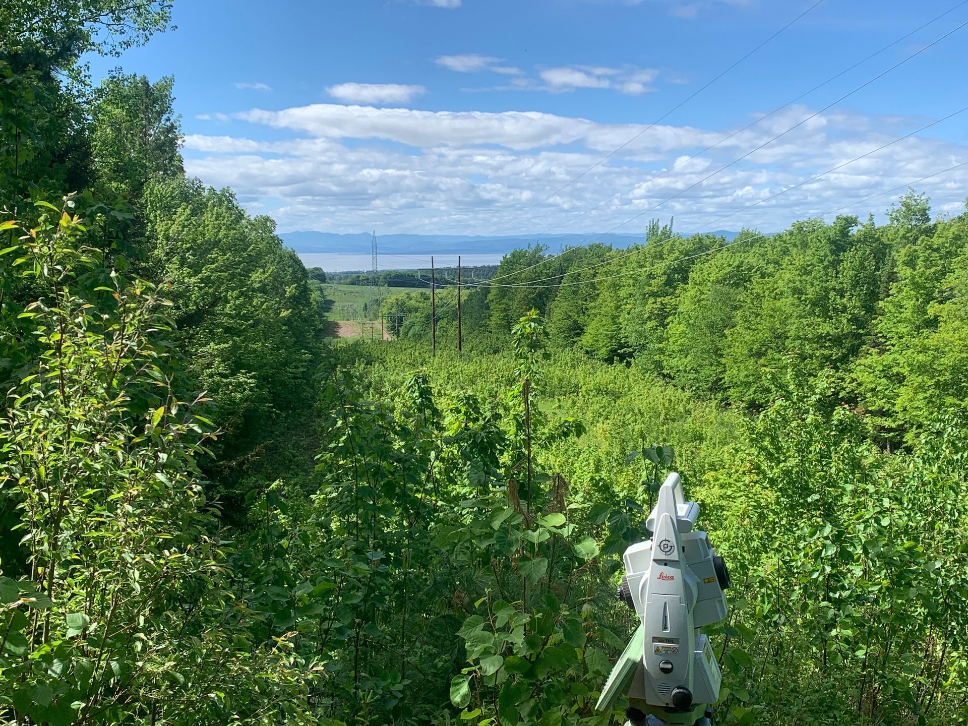 Au premier plan, une station totale de géomètre fait face à une clairière traversant une forêt luxuriante et verdoyante, en direction d'une baie lointaine.