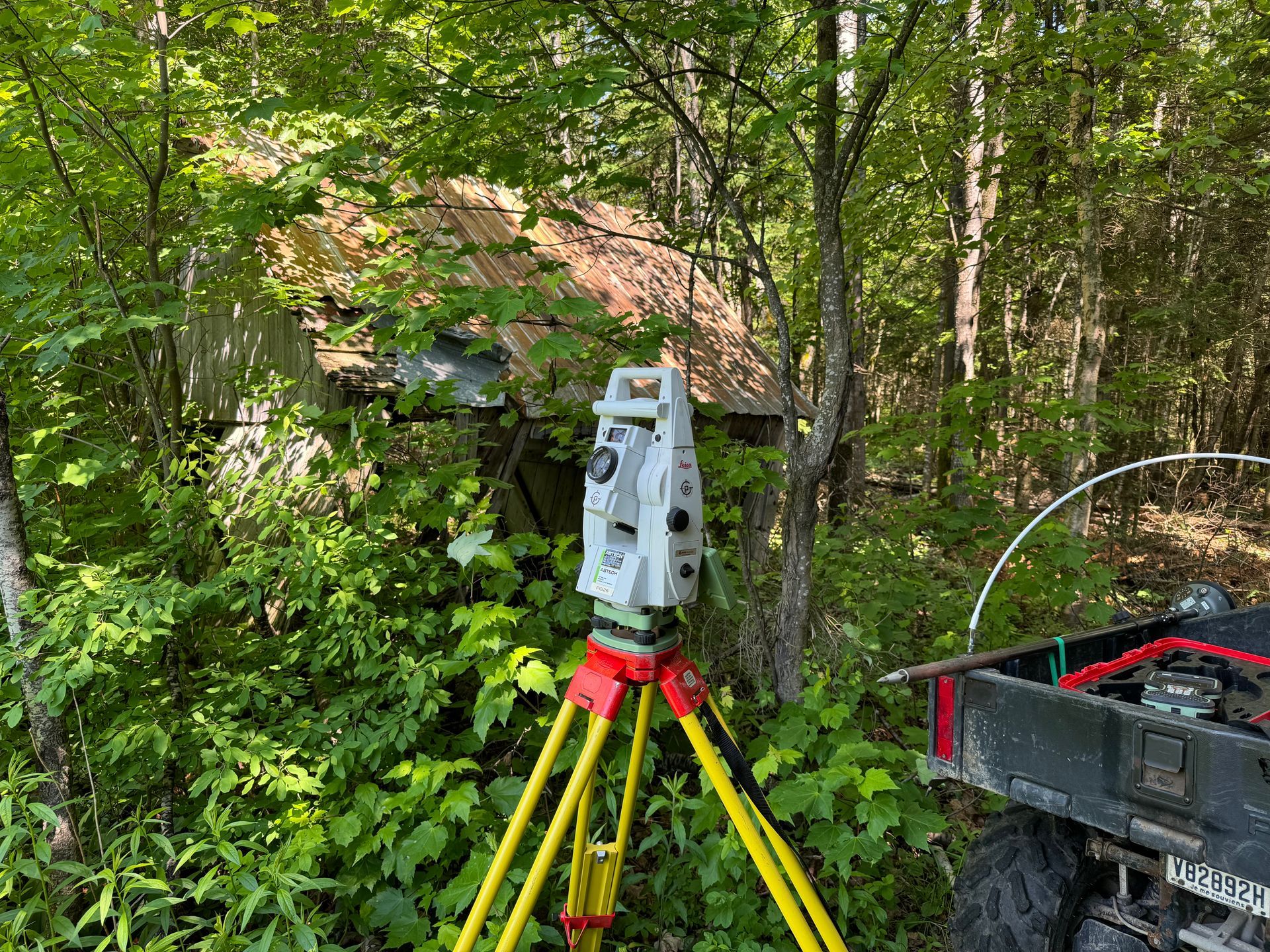 Un instrument d'arpentage gris se dresse sur un trépied jaune dans une forêt, près d'un véhicule et d'un petit bâtiment en bois usé par les intempéries.