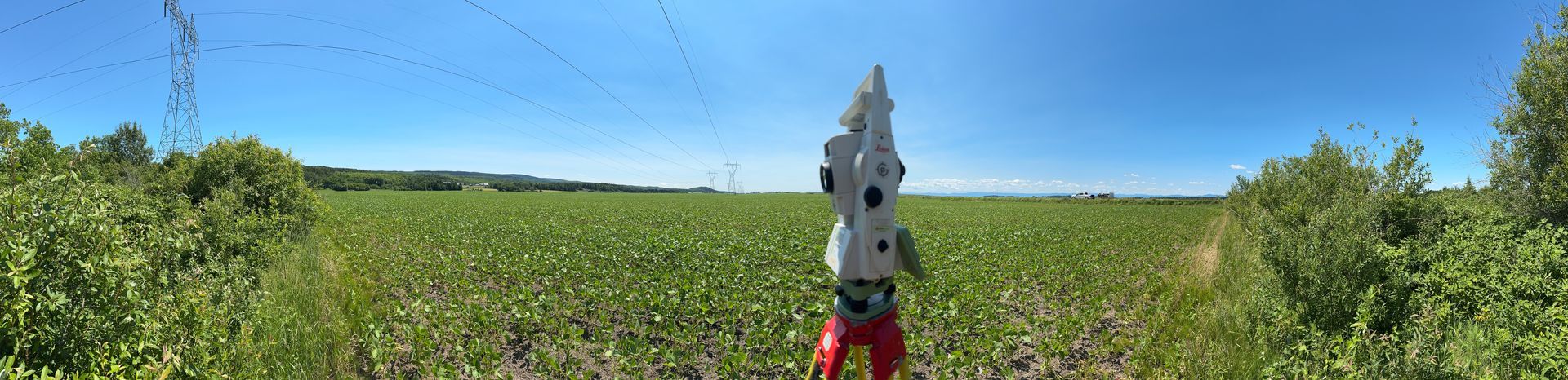 Un instrument de topographie est posé sur un trépied au milieu d'un vaste champ agricole ensoleillé, sous un ciel d'un bleu limpide.