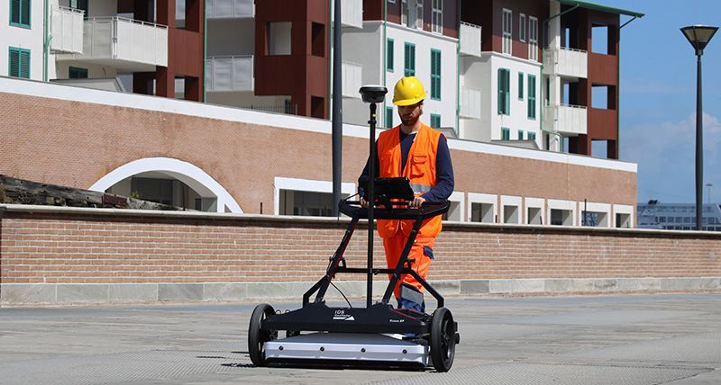Un ouvrier portant un équipement de sécurité pousse un radar de pénétration de sol sur une surface pavée devant un bâtiment.