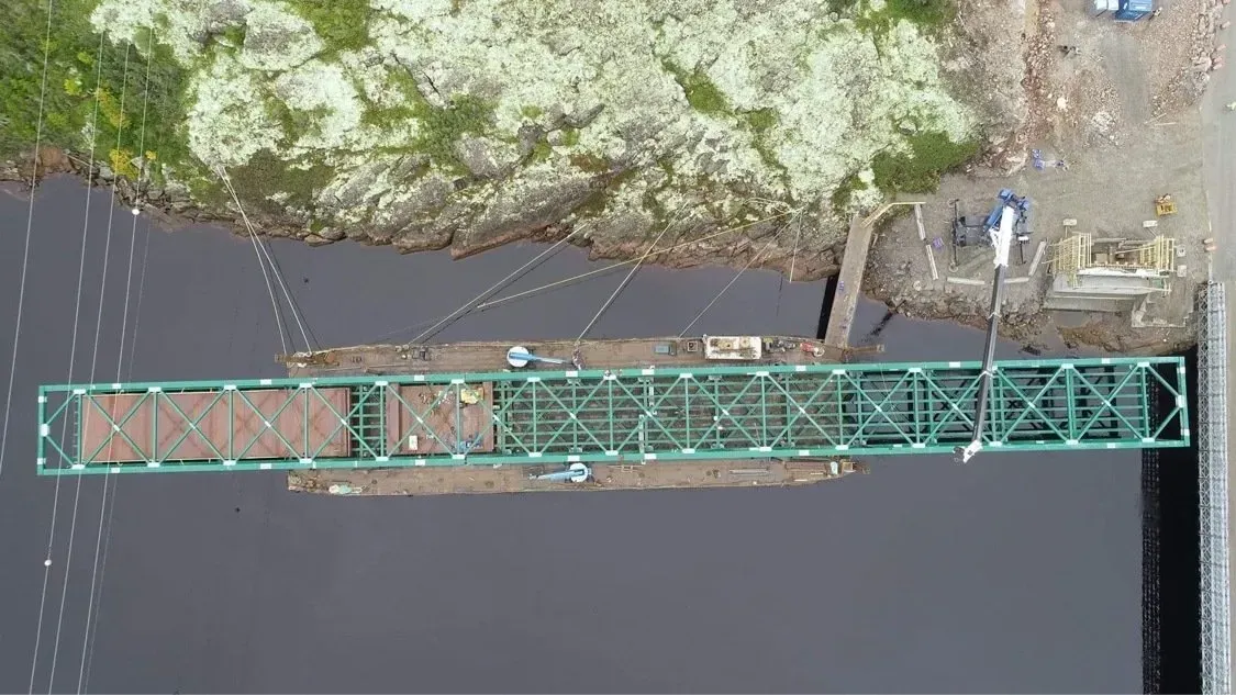 Vue aérienne d'une longue structure flottante en métal vert s'étendant d'un rivage rocheux vers une étendue d'eau sombre.