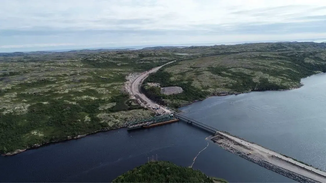 Vue aérienne d'un long et étroit pont traversant une étendue d'eau sombre, reliant deux zones de paysage rocheux et verdoyant.