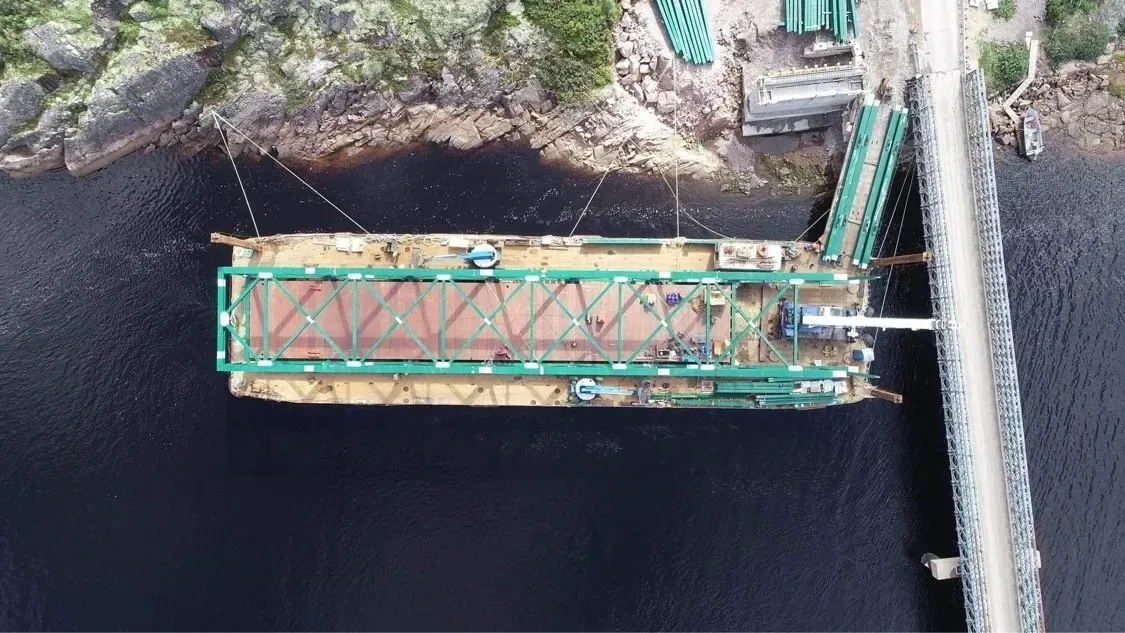Vue aérienne plongeante d'une barge à structure métallique verte amarrée à un quai en bois sur un lac sombre.