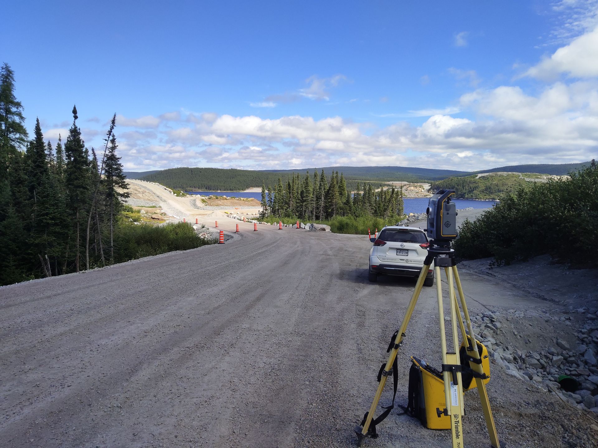 Un instrument de topographie sur trépied surplombe un chantier de construction de chemin de terre, avec une voiture garée à proximité et un lac en vue.