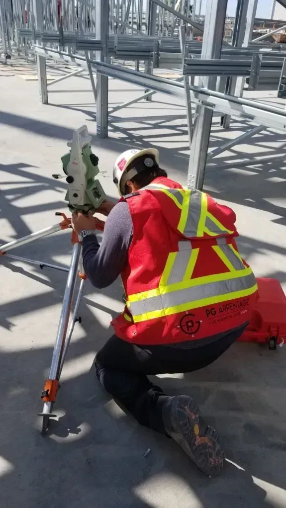 Un géomètre en construction, vêtu d'un gilet de sécurité rouge, est agenouillé sur un chantier en béton tout en réglant une station totale professionnelle.