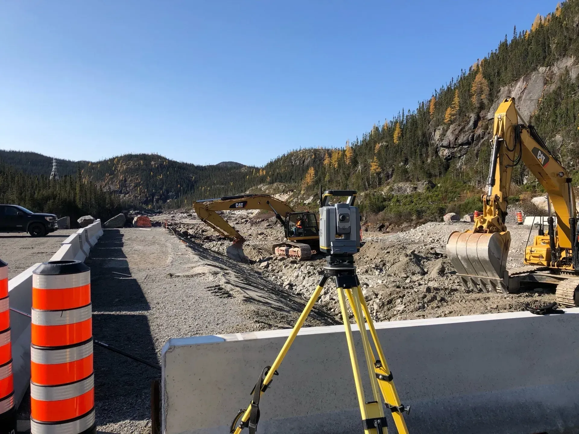 Un instrument de topographie sur trépied est installé sur un chantier de construction routière, à proximité d'excavatrices et de barils de sécurité orange.