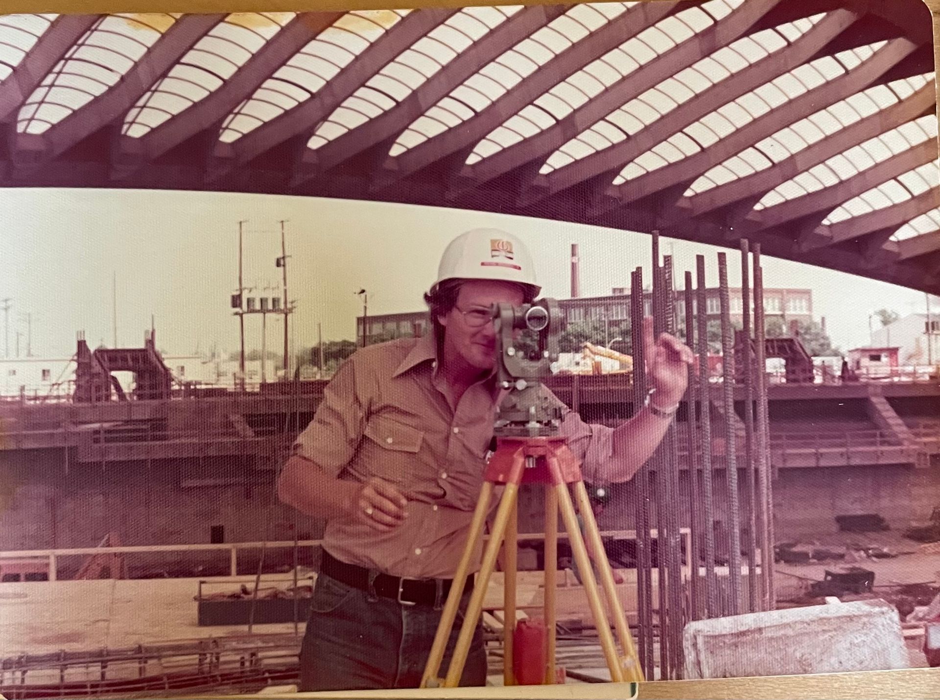 Sur un grand chantier de construction de stade, une personne portant un casque blanc utilise un théodolite sur trépied.