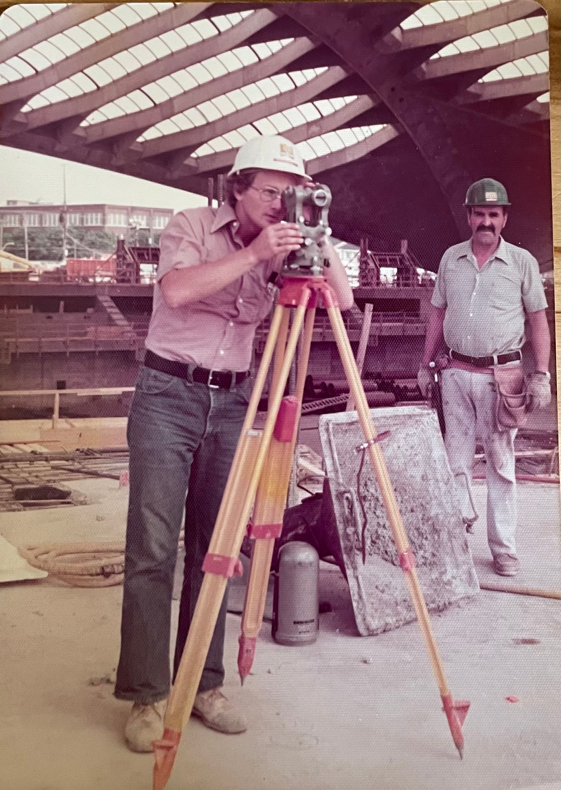 Deux ouvriers du bâtiment manipulent un théodolite sur un trépied en bois sous une grande structure en béton nervuré.