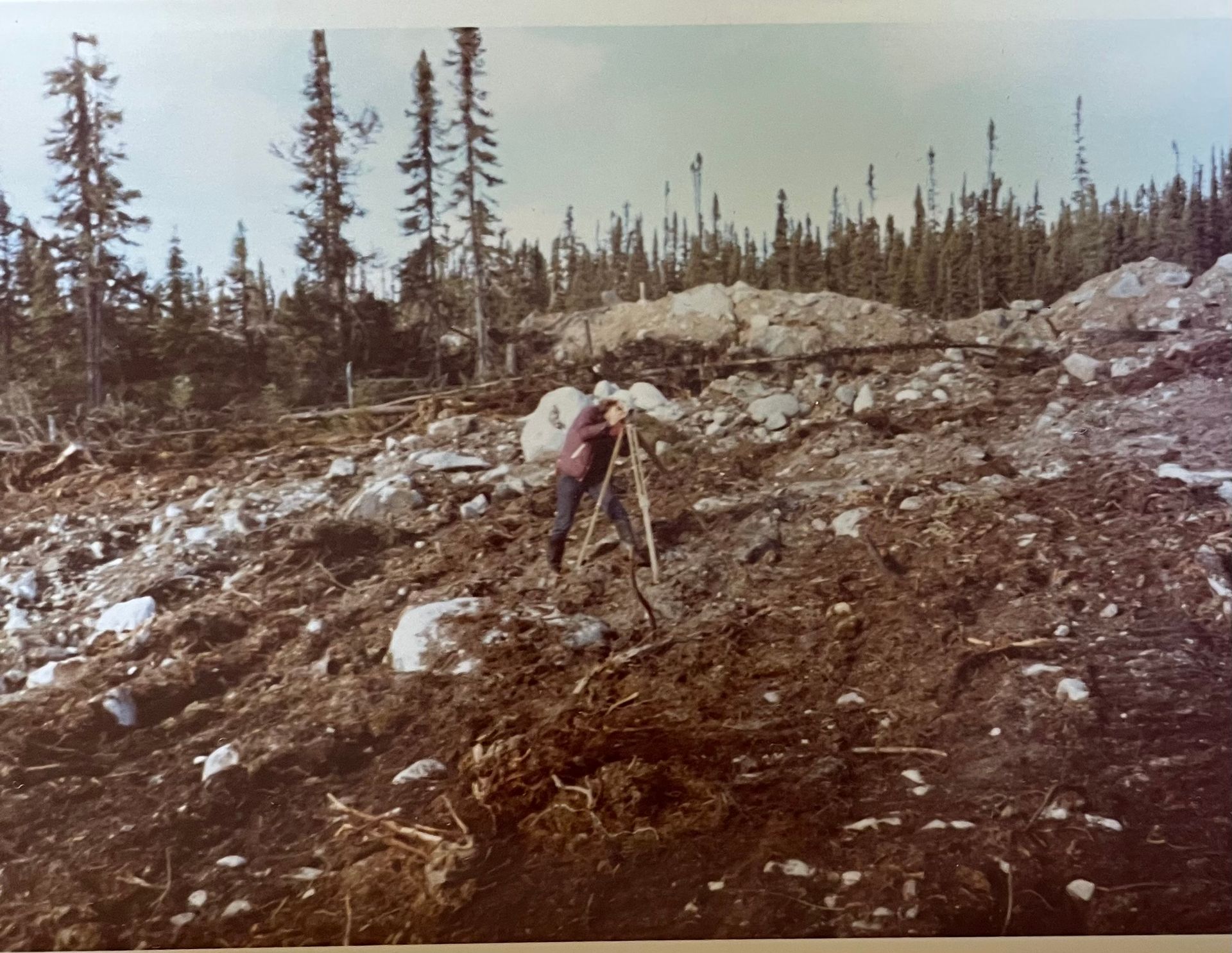 Une personne utilise un niveau de géomètre sur un versant de colline accidenté, rocailleux et boisé.