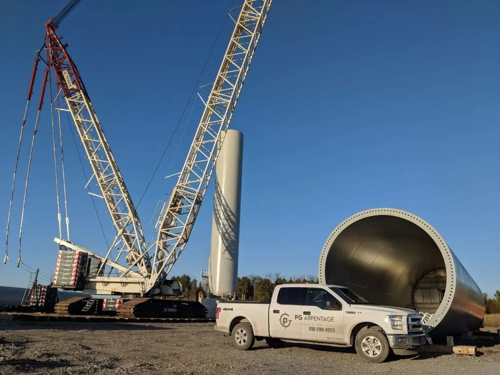 Une grande grue se dresse à côté d'une section verticale de tour d'éolienne et une camionnette est garée près de la base d'une tour horizontale.